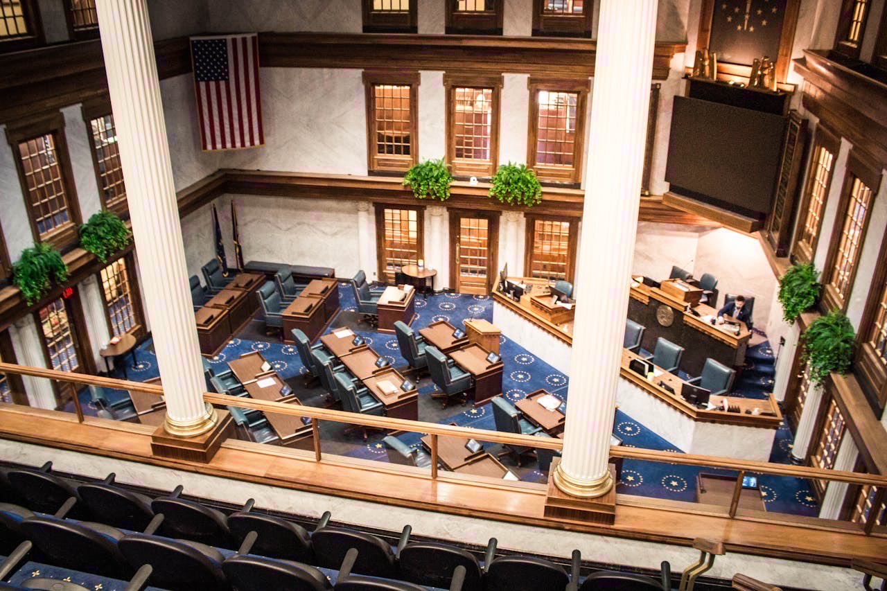 A state capitol building interior with legislative chamber desks and AI-themed documents, overhead shot, documentary style