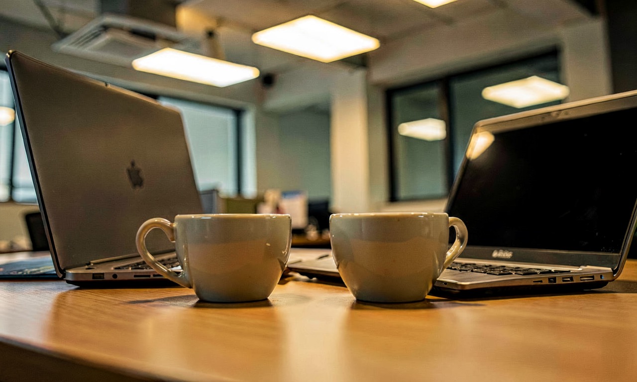 An office desk with a laptop showing an AI interface, two coffee cups, and a notebook, under soft overhead lighting.