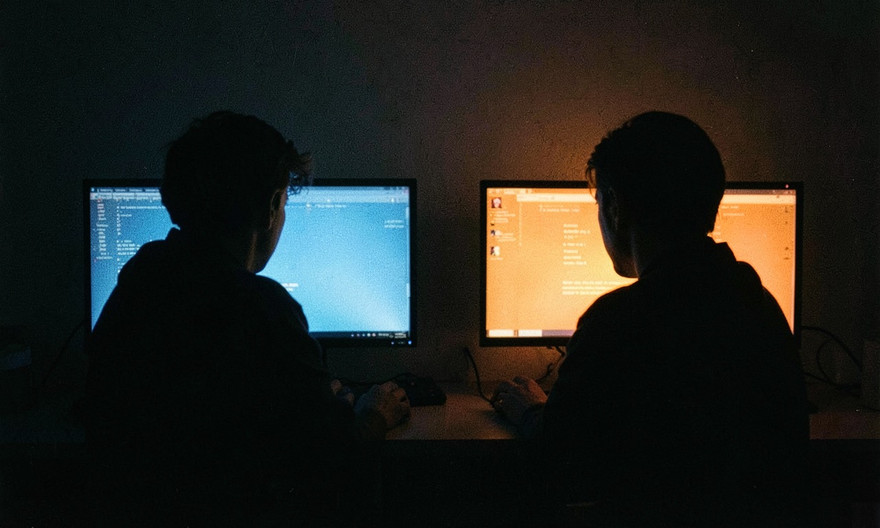 Two people silhouetted at computer screens, one real one artificial, a subtle glowing digital aura around one, dark room with blue screen light, cinematic portrait style