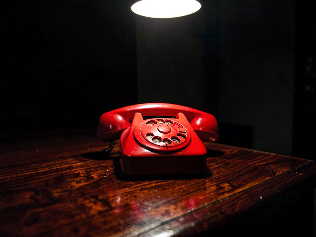 A red telephone sitting on a polished dark wood desk, single bright overhead light, no other objects, no visible text, no logos, no labels, dramatic chiaroscuro lighting, photorealistic