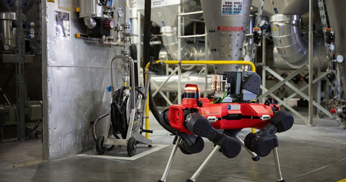 A red and black ANYmal quadruped robot inspecting an industrial plant with steel structures, silos and pipework around it