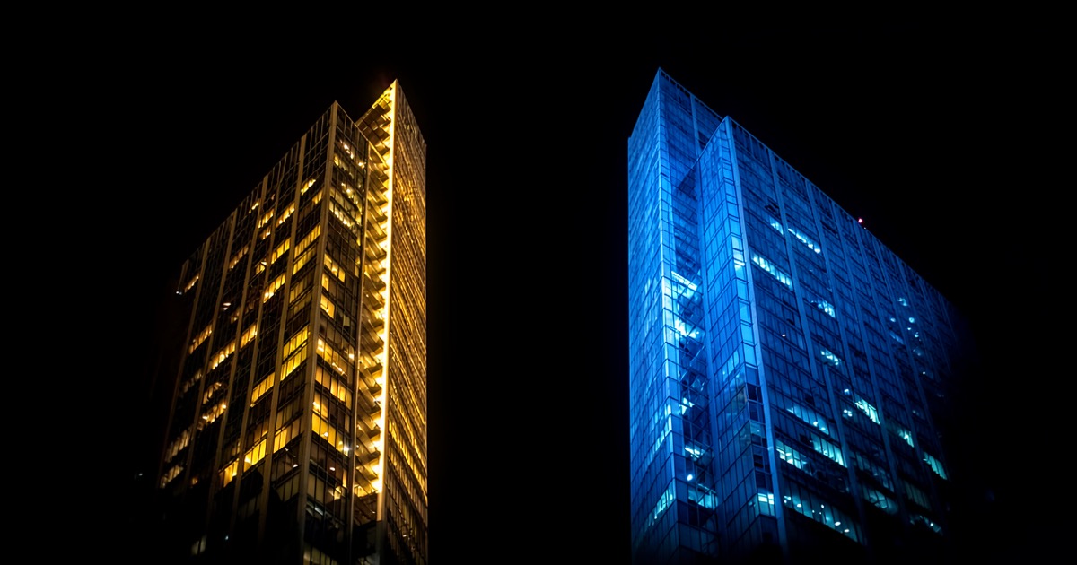 Two abstract corporate towers facing each other across a dark void, one glowing with warm light, the other with cool blue, suggesting a legal confrontation.