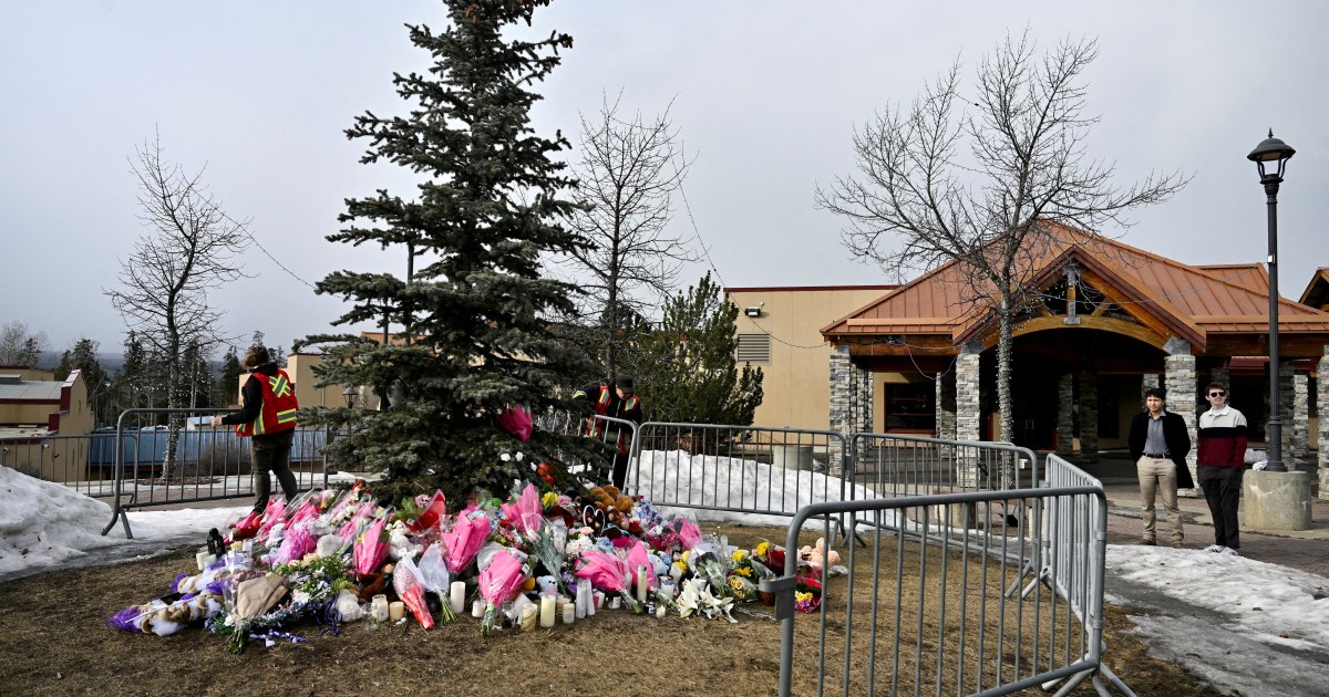 A memorial of flowers, candles and teddy bears around a spruce tree behind metal barricades outside a stone-and-timber school building under an overcast sky.