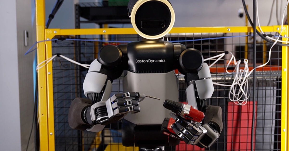 Atlas humanoid robot holding a red and black power drill in its four-fingered left hand and a drill bit in its right, inside a test lab