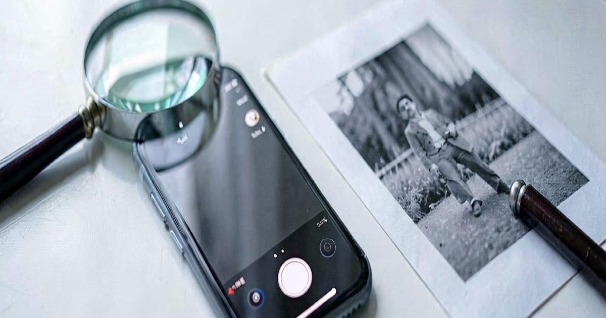 A smartphone camera app open on a desk next to a magnifying glass and a printed photograph, shot in natural light with shallow depth of field.