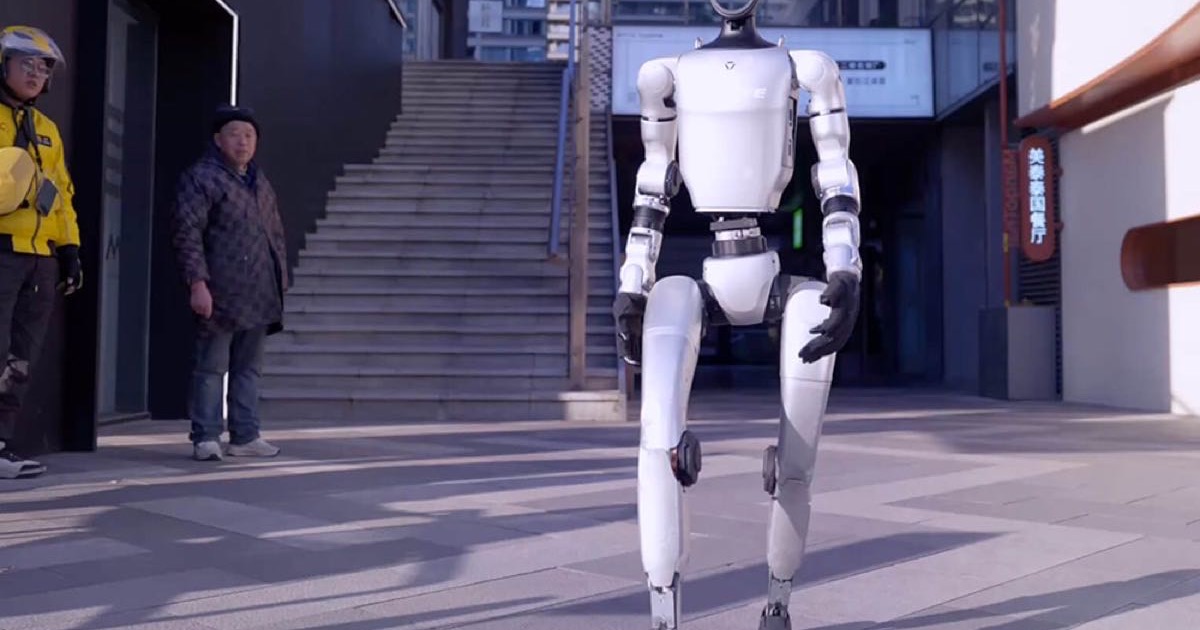 A white Unitree humanoid robot walking through a plaza outside an office building in China, a delivery rider and a man watching from the left