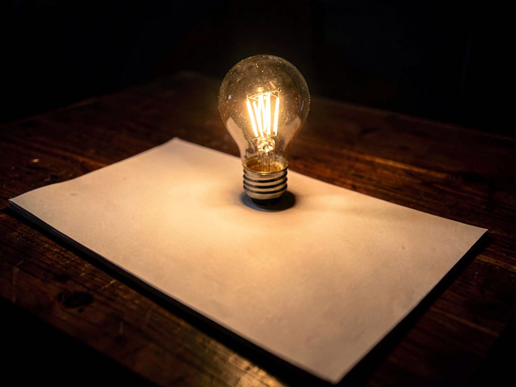 A single lit incandescent lightbulb casting warm glow over an open blank research paper on a dark wood desk, no visible text, no people, no logos, dramatic chiaroscuro lighting, photorealistic