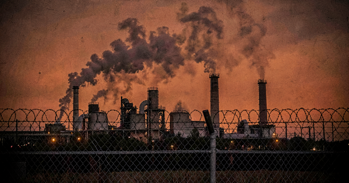 Industrial gas turbines at dusk with smoke plumes rising against an orange sky, viewed from behind a chain-link fence with barbed wire