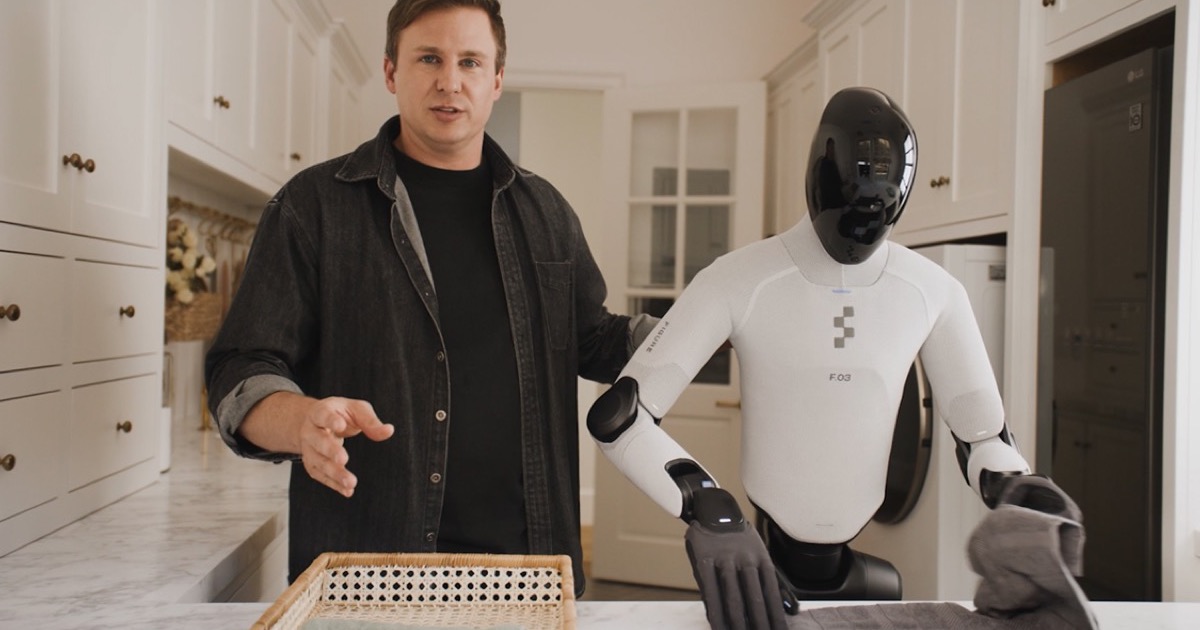 A Figure humanoid robot in a white bodysuit folds a towel at a kitchen counter while a man stands beside it in a bright home