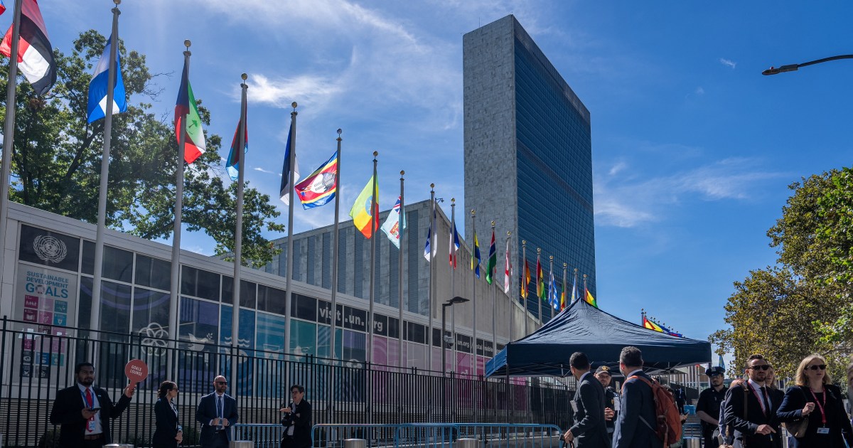 International flags on poles outside the United Nations headquarters in New York, with the UN Secretariat building rising behind a fence and visitors walking past on a sunny day.