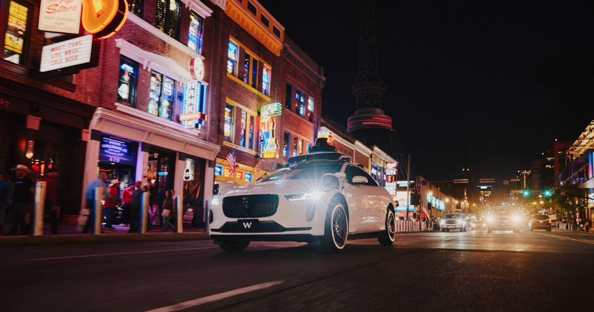 A white Waymo Jaguar I-Pace robotaxi with its lidar dome drives down Broadway past the neon honky-tonk signs of downtown Nashville at night
