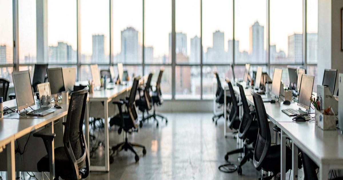 An empty open-plan office with rows of desks, chairs pushed in, soft afternoon light through floor-to-ceiling windows.