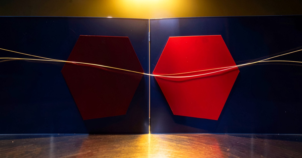 Abstract composition of a deep red geometric shape pressing against a navy blue barrier on a polished steel surface, with thin fibre-optic strands crossing the divide, warm golden lighting from above