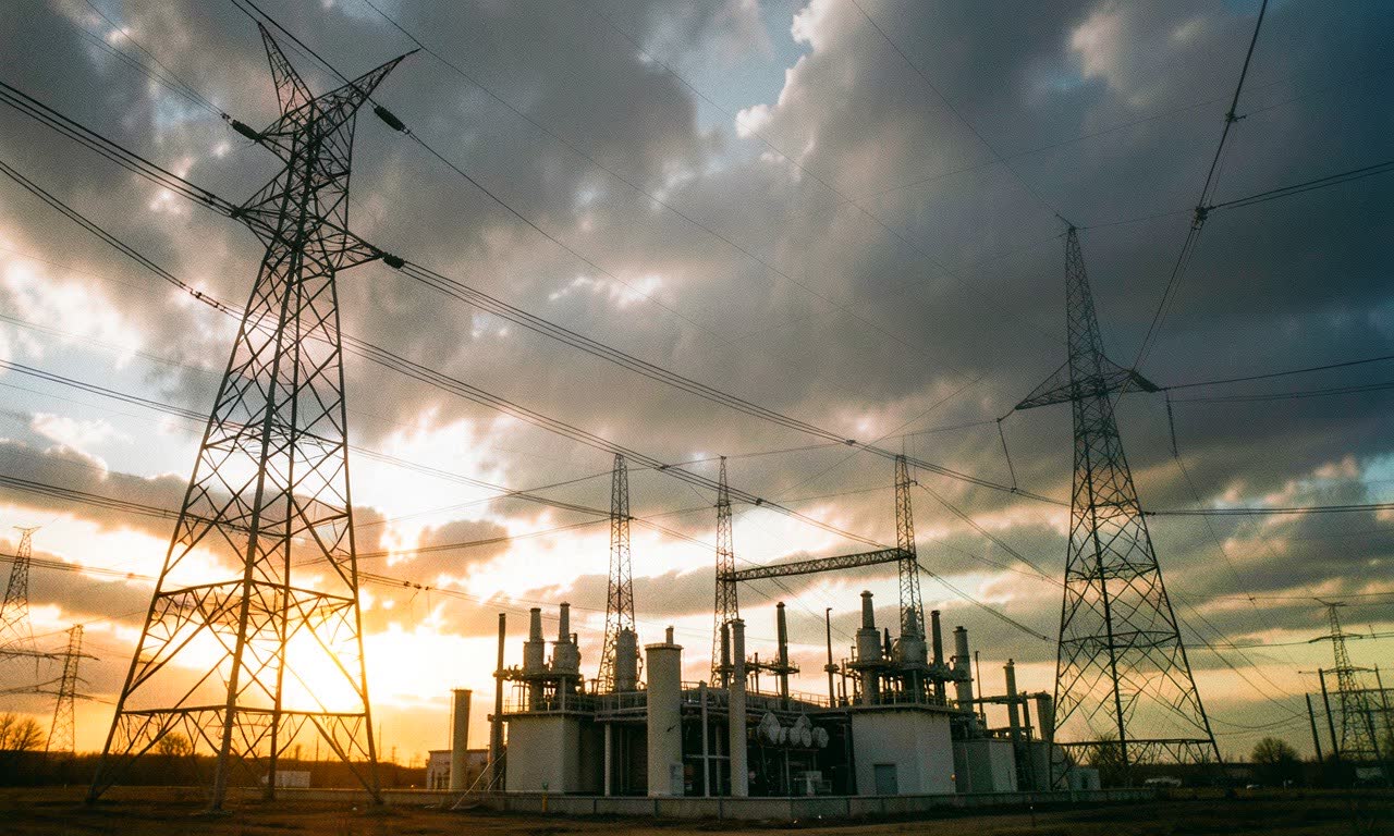 High voltage power transmission towers against cloudy sky, industrial electrical substation in foreground, documentary photography style