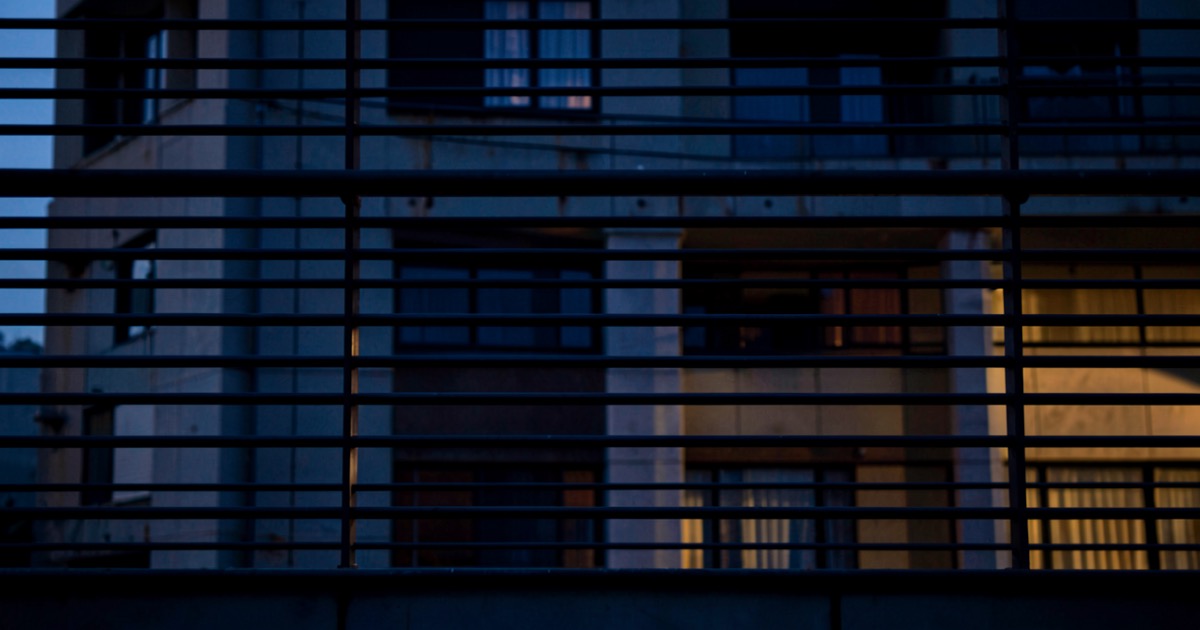 A dark government building facade at dusk, filtered through horizontal security bars casting long shadows