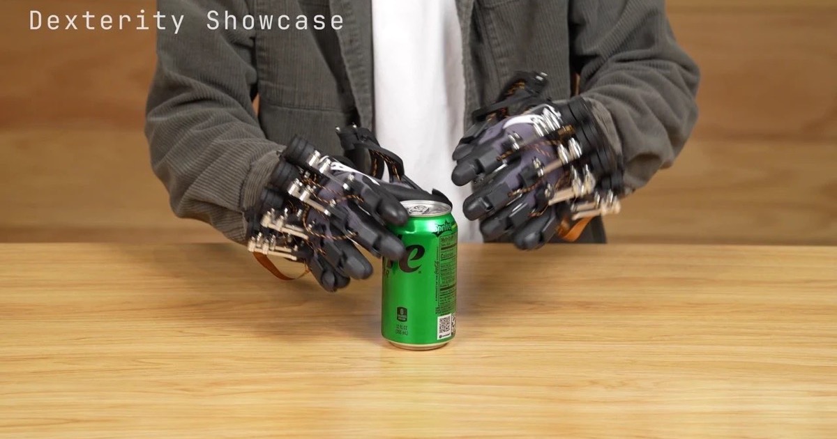 A person wearing sensor-equipped data gloves demonstrates fingertip dexterity over a beverage can on a table, in a still from an Origami Robotics dexterity showcase