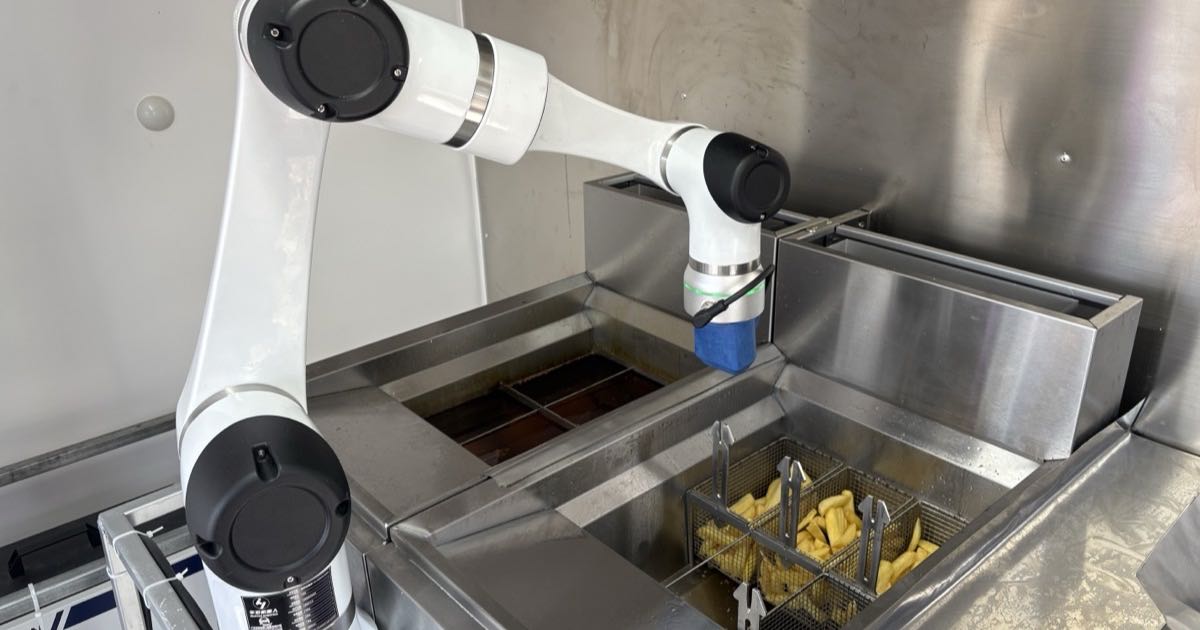 A white robotic arm holds a blue basket over a stainless steel deep fryer filled with hot oil, with a wire basket of cooked chips beside it.