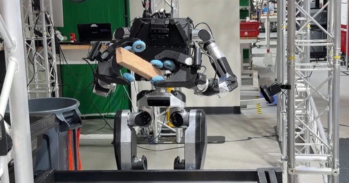 A metallic humanoid robot grips a length of timber while standing in a robotics laboratory workshop.