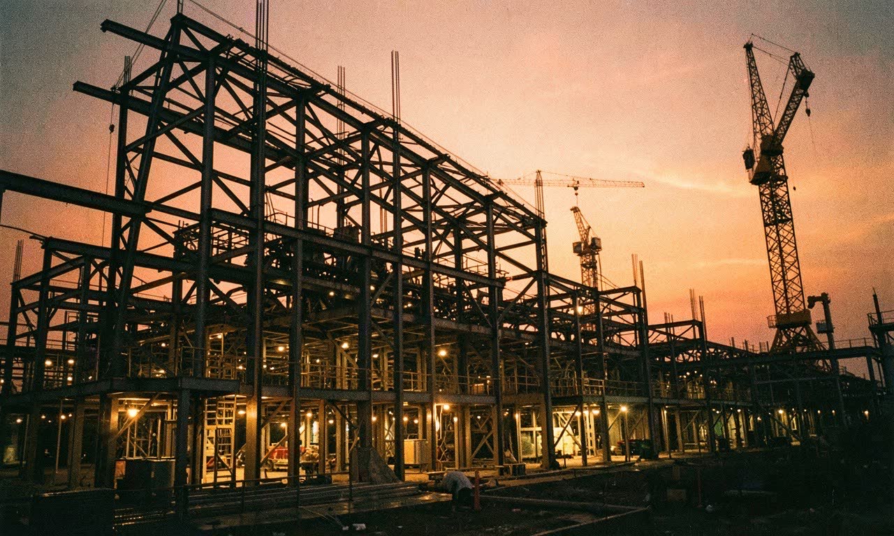 A semiconductor fabrication facility under construction at dusk, steel framework and crane silhouettes, warm industrial lighting