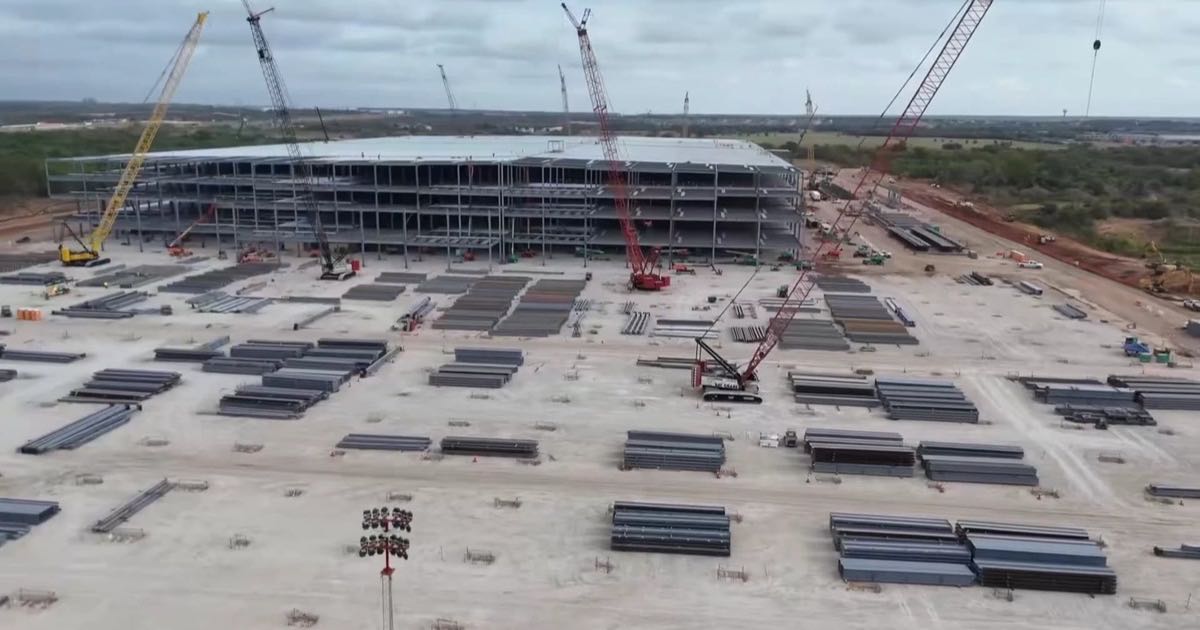 Aerial view of the Optimus factory construction site at Giga Texas, with cranes and a rising steel frame