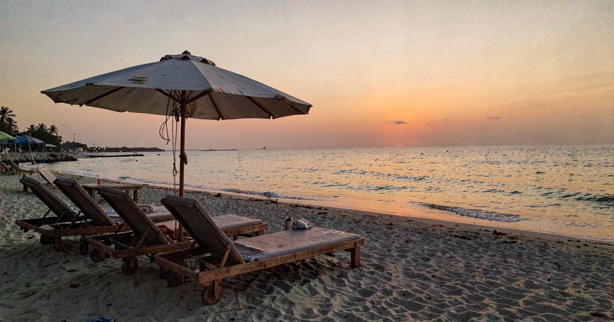 A beach resort at dusk with empty sun loungers and a faded umbrella, warm golden light on calm water