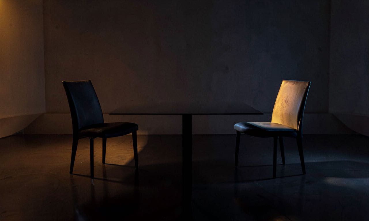 Two empty chairs facing each other across a dark polished table in a dimly lit room, symbolizing the diplomatic void between the White House and Anthropic's CEO.