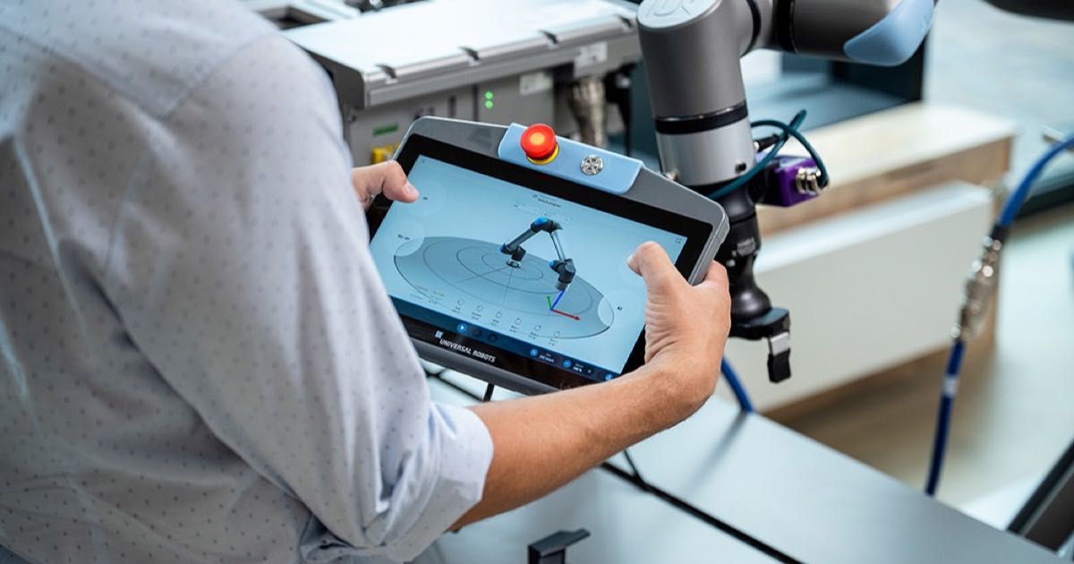 A technician programming a Universal Robots collaborative robot arm using the PolyScope teach pendant on a factory floor, the cobot arm visible beside the screen.